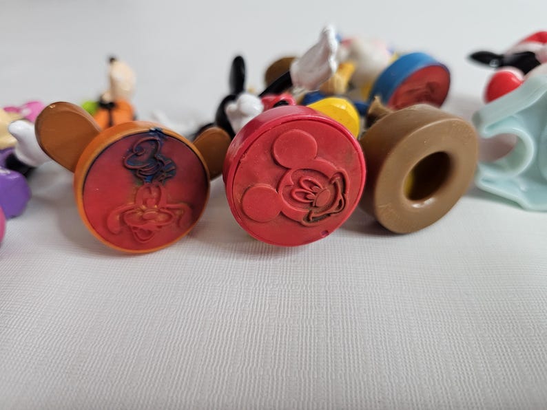 Op de afbeelding: Een verzameling kleurrijke plastic Disney-figuren met een rode, oranje en bruine basis. De figuren zijn Mickey Mouse, Donald Duck, Goofy en Pluto.