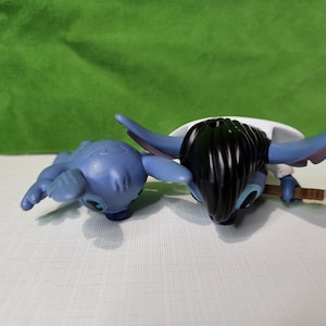 Peut inclure: Deux peluches bleues Stitch. Un Stitch est couch&eacute; sur le ventre, les bras tendus. L'autre Stitch est debout, les bras le long du corps, et a les cheveux noirs.
