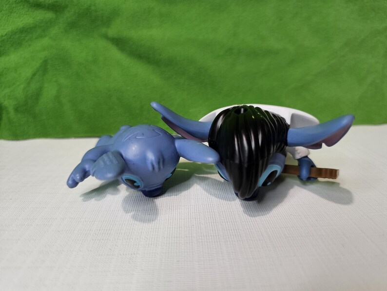 Peut inclure: Deux peluches bleues Stitch. Un Stitch est couch&eacute; sur le ventre, les bras tendus. L'autre Stitch est debout, les bras le long du corps, et a les cheveux noirs.