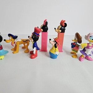 K&ouml;nnte beinhalten: Eine Sammlung von acht bunten Plastikfiguren von Disney-Charakteren. Zu den Figuren geh&ouml;ren Mickey Mouse, Minnie Mouse, Goofy, Pluto, Donald Duck, Daisy Duck und zwei weitere Figuren. Die Figuren sind alle in verschiedenen Posen und tragen verschiedene Outfits.