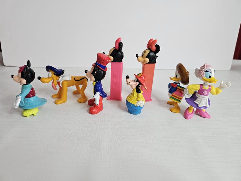 K&ouml;nnte beinhalten: Eine Sammlung von acht bunten Plastikfiguren von Disney-Charakteren. Zu den Figuren geh&ouml;ren Mickey Mouse, Minnie Mouse, Goofy, Pluto, Donald Duck, Daisy Duck und zwei weitere Figuren. Die Figuren sind alle in verschiedenen Posen und tragen verschiedene Outfits.