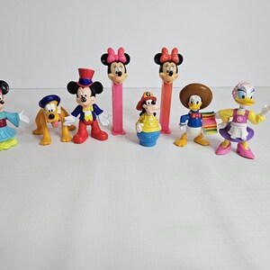 K&ouml;nnte beinhalten: Eine Sammlung von acht Disney-Figuren, darunter Mickey Mouse, Minnie Mouse, Donald Duck, Goofy und Pluto. Die Figuren sind aus Kunststoff und bunt. Sie stehen alle auf einer wei&szlig;en Oberfl&auml;che.