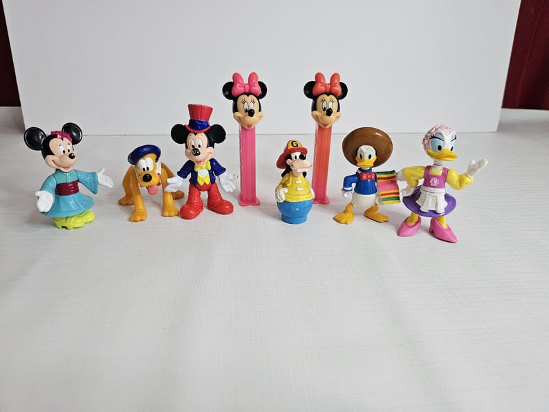 K&ouml;nnte beinhalten: Eine Sammlung von acht Disney-Figuren, darunter Mickey Mouse, Minnie Mouse, Donald Duck, Goofy und Pluto. Die Figuren sind aus Kunststoff und bunt. Sie stehen alle auf einer wei&szlig;en Oberfl&auml;che.