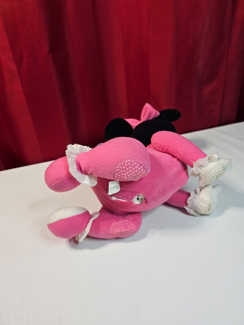 K&ouml;nnte beinhalten: Rosa Pl&uuml;schtier mit schwarzen Ohren und einer wei&szlig;en Schleife. Das Spielzeug liegt auf dem R&uuml;cken mit den Beinen in der Luft. Das Spielzeug hat einen wei&szlig;en Fleck auf der Brust und einen wei&szlig;en Fleck an jedem Fu&szlig;.