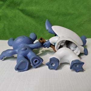 Peut inclure: Deux figurines en plastique bleues et blanches. La figurine bleue est allong&eacute;e sur le dos, les bras tendus. La figurine blanche est debout, les jambes &eacute;cart&eacute;es et les bras lev&eacute;s. Les deux figurines ont une apparence de dessin anim&eacute;.