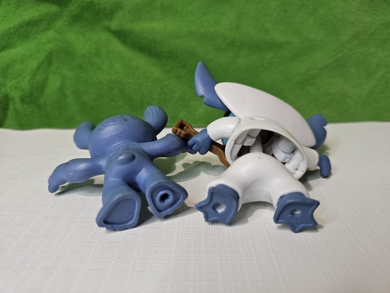 Peut inclure: Deux figurines en plastique bleues et blanches. La figurine bleue est allong&eacute;e sur le dos, les bras tendus. La figurine blanche est debout, les jambes &eacute;cart&eacute;es et les bras lev&eacute;s. Les deux figurines ont une apparence de dessin anim&eacute;.