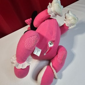 K&ouml;nnte beinhalten: Rosa Pl&uuml;schtier mit wei&szlig;en Akzenten und einer schwarzen Schleife. Das Spielzeug hat ein Etikett auf der R&uuml;ckseite mit der Aufschrift "Hergestellt in China, Ganz neues Material, Reg. Nr. PA 12002 (CN)  ", und "The Manhattan Toy Company".
