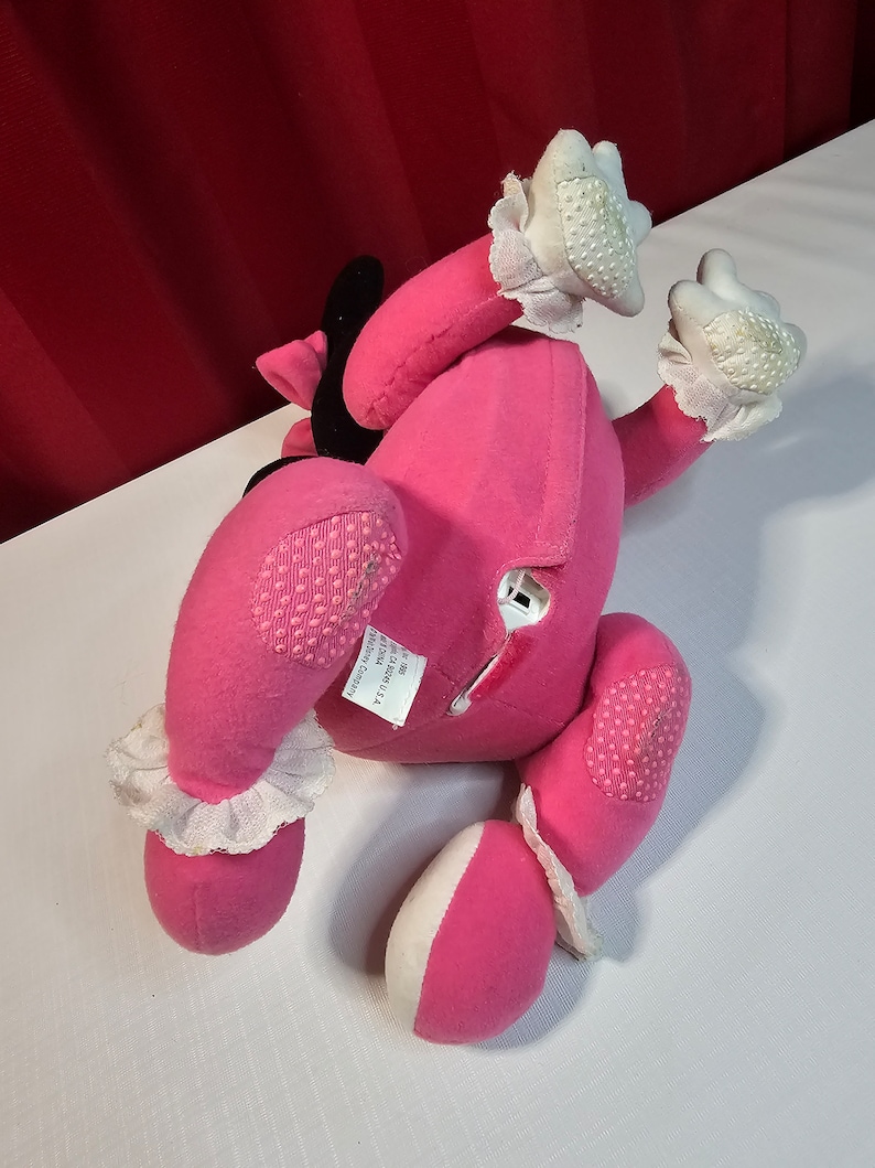 K&ouml;nnte beinhalten: Rosa Pl&uuml;schtier mit wei&szlig;en Akzenten und einer schwarzen Schleife. Das Spielzeug hat ein Etikett auf der R&uuml;ckseite mit der Aufschrift "Hergestellt in China, Ganz neues Material, Reg. Nr. PA 12002 (CN)  ", und "The Manhattan Toy Company".