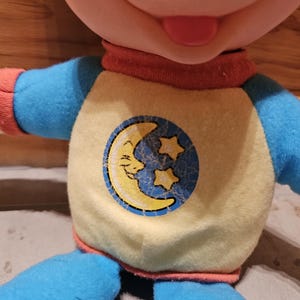 May include: A plush doll with a smiling face and a red tongue. The doll wears a yellow shirt with a blue circle featuring a crescent moon and stars. The doll's sleeves and pants are blue, and the collar is red.