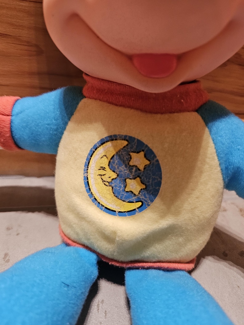 May include: A plush doll with a smiling face and a red tongue. The doll wears a yellow shirt with a blue circle featuring a crescent moon and stars. The doll's sleeves and pants are blue, and the collar is red.