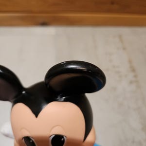 May include: A close-up of a Mickey Mouse figurine. The figurine has a black head and ears, a tan face, and large black eyes. The figure is wearing a blue shirt with red and yellow accents.