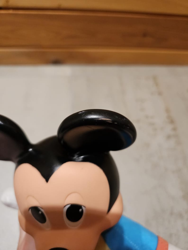 May include: A close-up of a Mickey Mouse figurine. The figurine has a black head and ears, a tan face, and large black eyes. The figure is wearing a blue shirt with red and yellow accents.