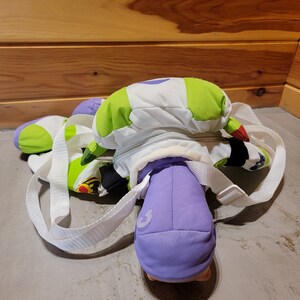 May include: A green, white, and purple plush backpack with a zippered pocket and adjustable straps. The backpack is shaped like a character from a popular animated film.