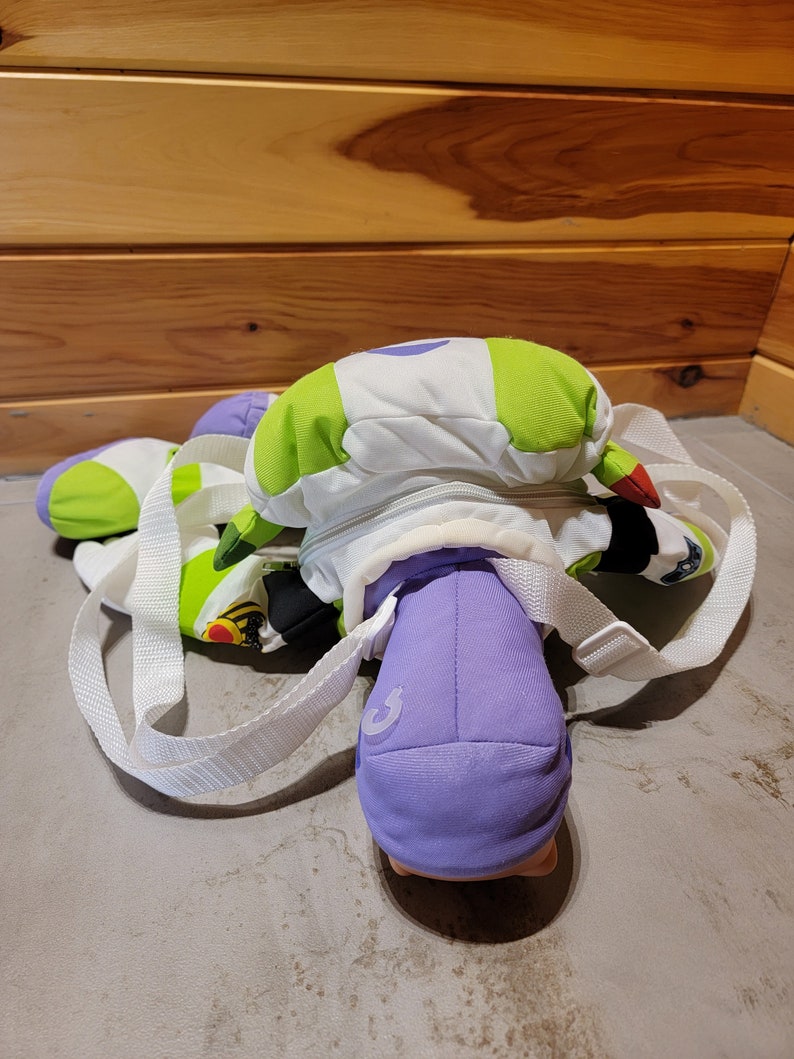 May include: A green, white, and purple plush backpack with a zippered pocket and adjustable straps. The backpack is shaped like a character from a popular animated film.