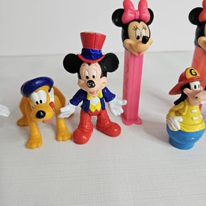 K&ouml;nnte beinhalten: Eine Sammlung von Vintage-Disney-Figuren, darunter Mickey Mouse, Minnie Mouse, Goofy und Pluto. Die Figuren sind aus Kunststoff und in leuchtenden Farben gehalten. Mickey Mouse tr&auml;gt ein rot-wei&szlig;-blaues Outfit, Minnie Mouse ein rosa Kleid und eine Schleife, Goofy eine gelbe Feuerwehruniform und Pluto einen blauen Hut.