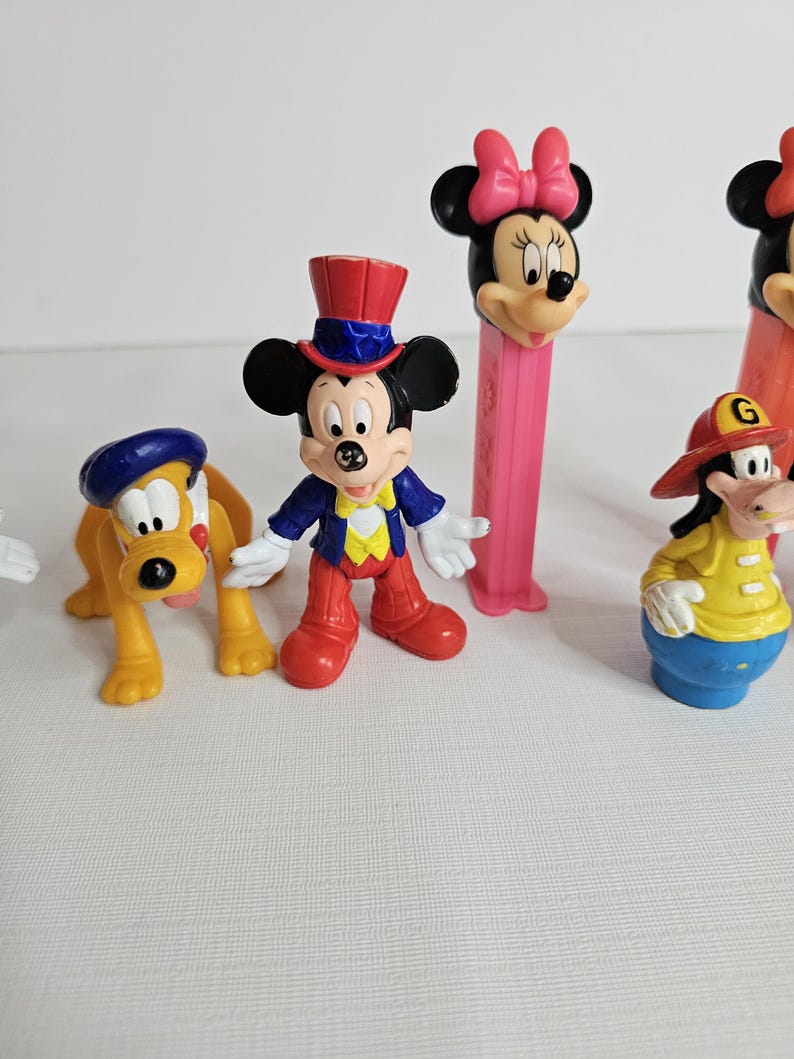 K&ouml;nnte beinhalten: Eine Sammlung von Vintage-Disney-Figuren, darunter Mickey Mouse, Minnie Mouse, Goofy und Pluto. Die Figuren sind aus Kunststoff und in leuchtenden Farben gehalten. Mickey Mouse tr&auml;gt ein rot-wei&szlig;-blaues Outfit, Minnie Mouse ein rosa Kleid und eine Schleife, Goofy eine gelbe Feuerwehruniform und Pluto einen blauen Hut.