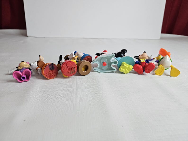 Op de afbeelding: Een verzameling van negen kleine plastic Disney-figuren. De figuren omvatten Mickey Mouse, Minnie Mouse, Donald Duck, Goofy en Pluto. De figuren staan allemaal in verschillende poses en zijn felgekleurd.
