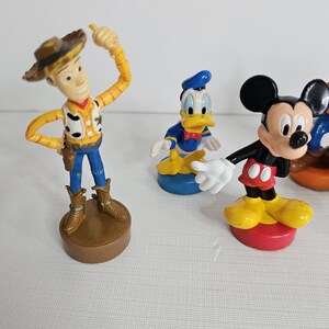Op de afbeelding: Een set van vijf plastic figuren. De figuren zijn Woody uit Toy Story, Mickey Mouse, Donald Duck, Goofy en Pluto. De figuren staan allemaal op kleine, kleurrijke voetstukken.