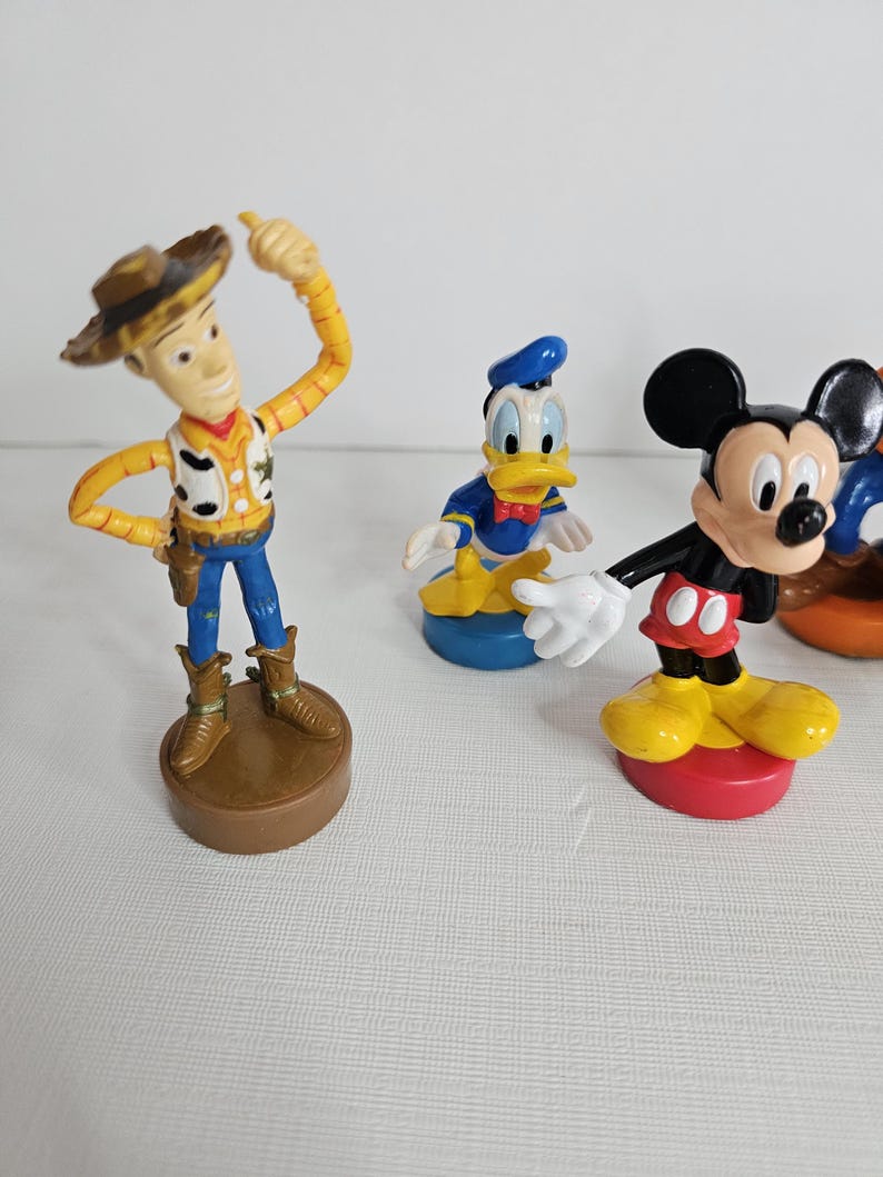Op de afbeelding: Een set van vijf plastic figuren. De figuren zijn Woody uit Toy Story, Mickey Mouse, Donald Duck, Goofy en Pluto. De figuren staan allemaal op kleine, kleurrijke voetstukken.