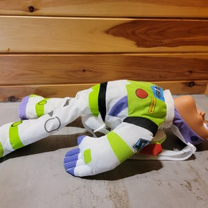 May include: A plush toy of Buzz Lightyear from the Toy Story franchise. The toy is lying on its back with its arms and legs outstretched. The toy is wearing a white and green spacesuit with purple accents. The toy has a red button on its chest and the text "LIGHTYEAR" is printed on its chest.