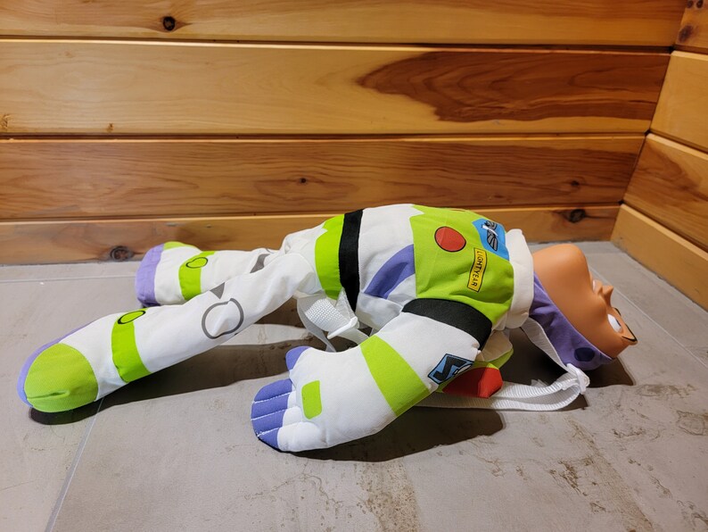 May include: A plush toy of Buzz Lightyear from the Toy Story franchise. The toy is lying on its back with its arms and legs outstretched. The toy is wearing a white and green spacesuit with purple accents. The toy has a red button on its chest and the text "LIGHTYEAR" is printed on its chest.