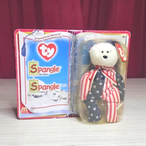 Peut inclure: Un ours en peluche Ty Beanie Baby nommé Spangle the Bear, dans son emballage d'origine. L'ours a une tête blanche et un corps avec un motif de drapeau américain rouge, blanc et bleu. L'emballage comprend le texte "International Bears II" et "Spangle the Bear."