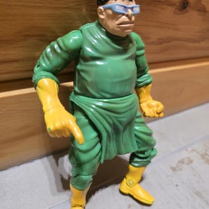 May include: A Dr. Octopus action figure. The figure is wearing a green outfit with yellow gloves and boots. The figure has a bald head with black hair and blue-tinted glasses. The figure is in a pointing pose.