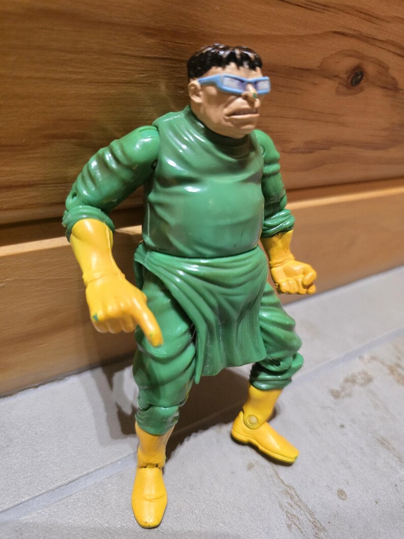 May include: A Dr. Octopus action figure. The figure is wearing a green outfit with yellow gloves and boots. The figure has a bald head with black hair and blue-tinted glasses. The figure is in a pointing pose.