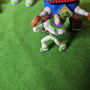 May include: A green, white and purple plastic toy figure of Buzz Lightyear from the Toy Story franchise. The figure is standing on a green surface and is holding a blaster in his right hand.
