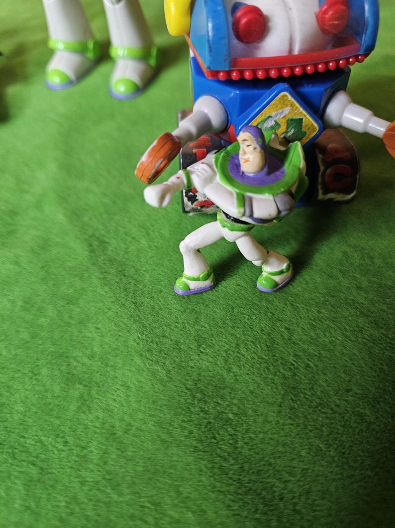 May include: A green, white and purple plastic toy figure of Buzz Lightyear from the Toy Story franchise. The figure is standing on a green surface and is holding a blaster in his right hand.