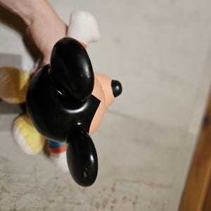 May include: A Mickey Mouse figurine with black ears, a black head, and a tan face. The figurine has a white glove and a red, blue, and white outfit. The figurine is being held by a person.