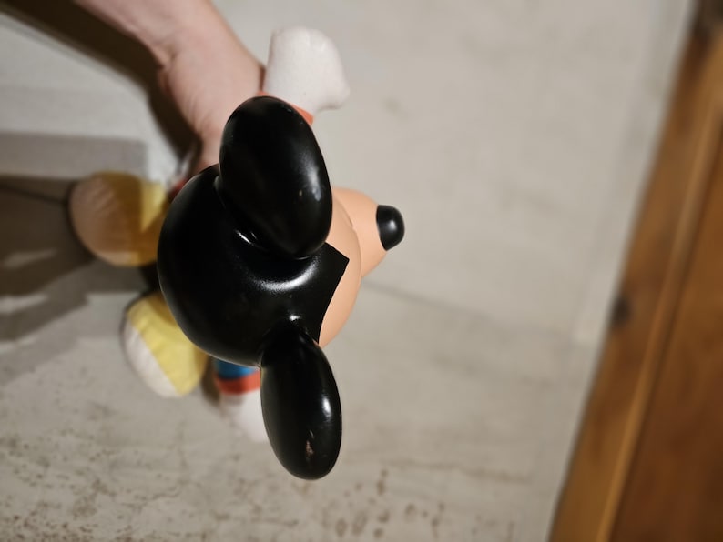 May include: A Mickey Mouse figurine with black ears, a black head, and a tan face. The figurine has a white glove and a red, blue, and white outfit. The figurine is being held by a person.