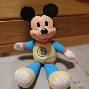 May include: A plush Mickey Mouse doll with black ears, a pink face, and a blue and yellow pajama set. The pajamas feature a moon and stars design. The doll has white hands and feet with a dotted pattern.