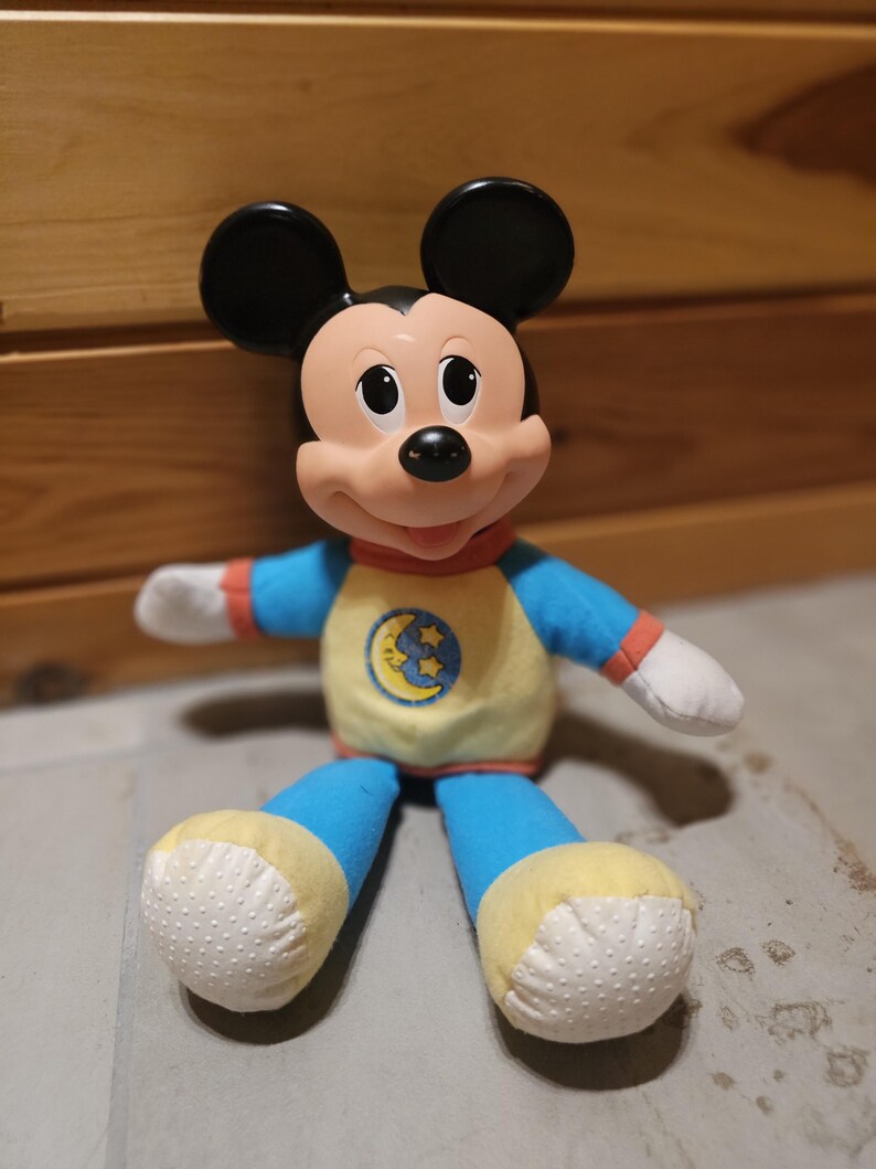 May include: A plush Mickey Mouse doll with black ears, a pink face, and a blue and yellow pajama set. The pajamas feature a moon and stars design. The doll has white hands and feet with a dotted pattern.