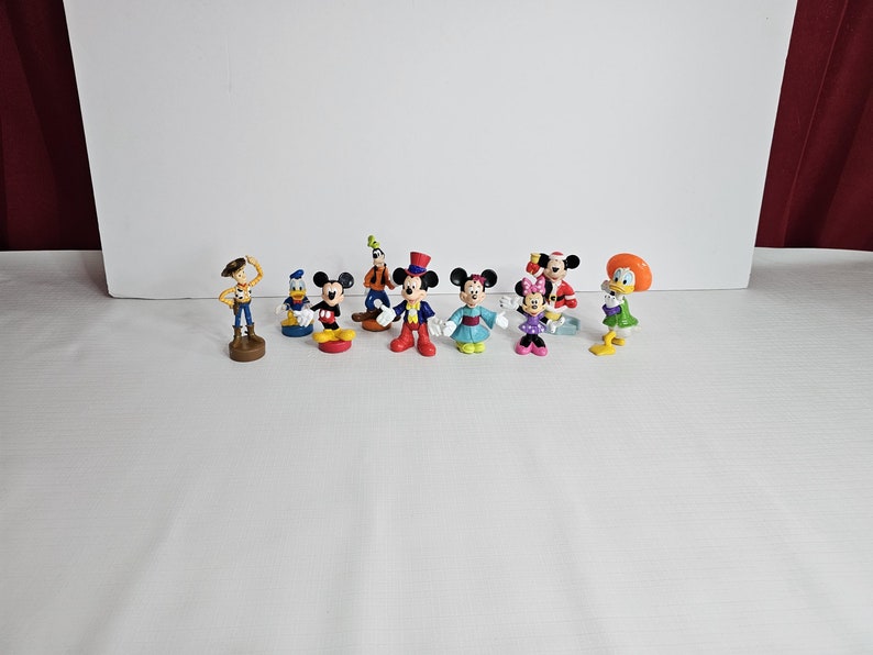Op de afbeelding: Een verzameling van negen kleine plastic figuurtjes van populaire Disney-personages, waaronder Mickey Mouse, Minnie Mouse, Donald Duck, Goofy en Woody uit Toy Story. De figuurtjes staan allemaal in verschillende poses en zijn felgekleurd.