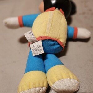 May include: A vintage plush toy featuring a black head, blue body, yellow accents, and white hands and feet. The toy has a red trim and a tag attached to the back. The toy is in a seated position.