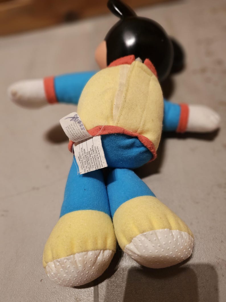 May include: A vintage plush toy featuring a black head, blue body, yellow accents, and white hands and feet. The toy has a red trim and a tag attached to the back. The toy is in a seated position.