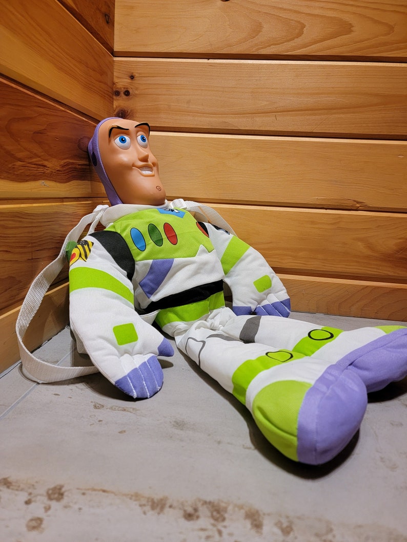 May include: A plush Buzz Lightyear toy from the Toy Story franchise. The toy is wearing a green and white spacesuit with a purple chest piece and a large, cartoonish face. The toy is sitting on a light gray surface.
