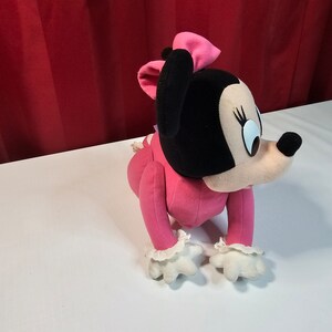 K&ouml;nnte beinhalten: Rosa Pl&uuml;sch-Minnie Mouse Spielzeug mit schwarzer Nase und gro&szlig;en Augen. Sie tr&auml;gt ein rosa Kleid mit wei&szlig;en Spitzenbesatz und einer rosa Schleife.
