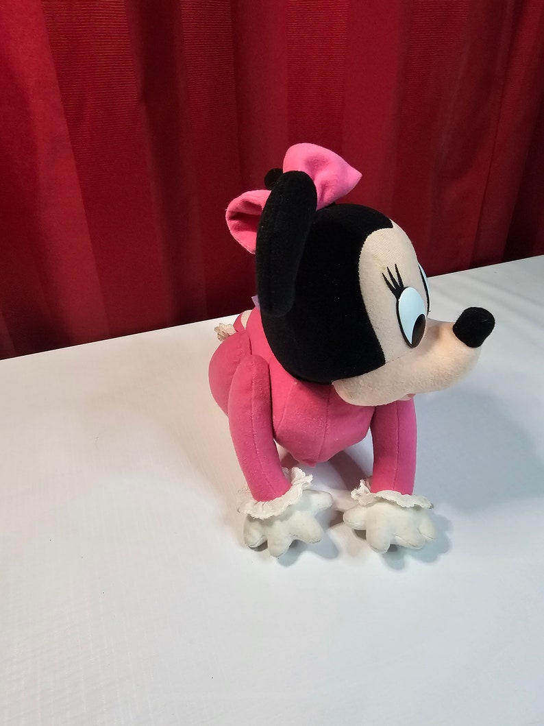 K&ouml;nnte beinhalten: Rosa Pl&uuml;sch-Minnie Mouse Spielzeug mit schwarzer Nase und gro&szlig;en Augen. Sie tr&auml;gt ein rosa Kleid mit wei&szlig;en Spitzenbesatz und einer rosa Schleife.