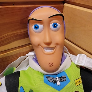May include: A plush toy of Buzz Lightyear from the Toy Story franchise. The toy is wearing his signature space ranger suit with a green vest, blue and white wings, and a yellow patch that says "LIGHT YEAR". The toy has a purple headband and a detailed face with blue eyes.