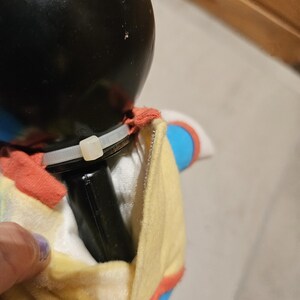 May include: A black plush toy with a black head and a yellow and red outfit. The toy has a black rectangular object attached to it with a white zip tie.