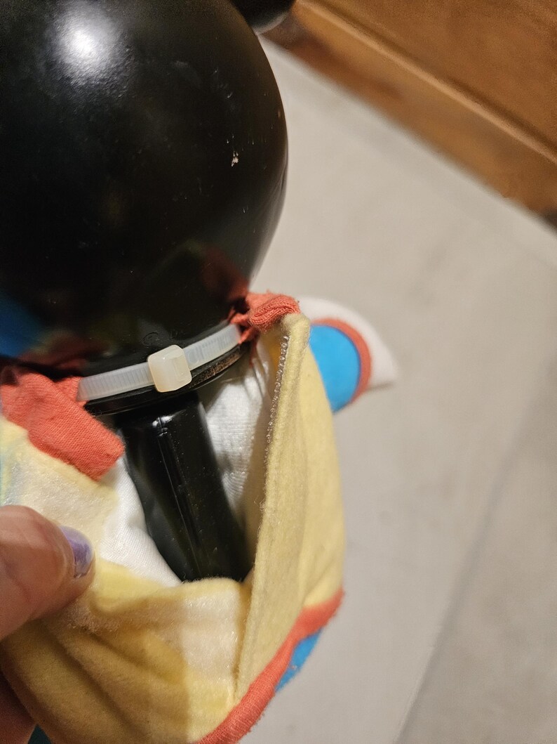 May include: A black plush toy with a black head and a yellow and red outfit. The toy has a black rectangular object attached to it with a white zip tie.
