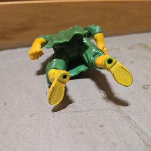 May include: A green and yellow action figure, lying on a gray surface. The figure has yellow arms and feet, and green clothing. The figure's feet are pointing towards the viewer.