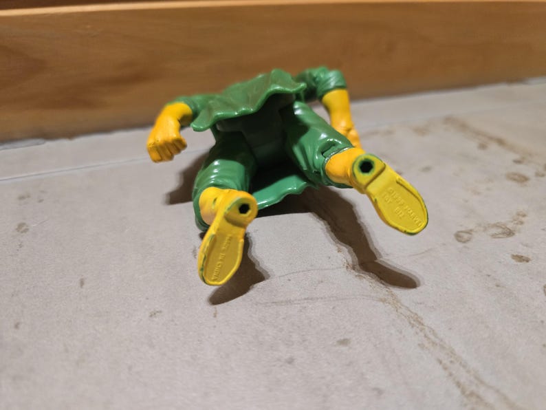 May include: A green and yellow action figure, lying on a gray surface. The figure has yellow arms and feet, and green clothing. The figure's feet are pointing towards the viewer.