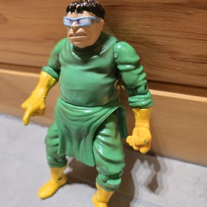 May include: A vintage action figure of a character in a green outfit with yellow gloves and boots. The figure has a stern expression, wearing blue-rimmed glasses. The figure is posed in a dynamic stance, suggesting action.
