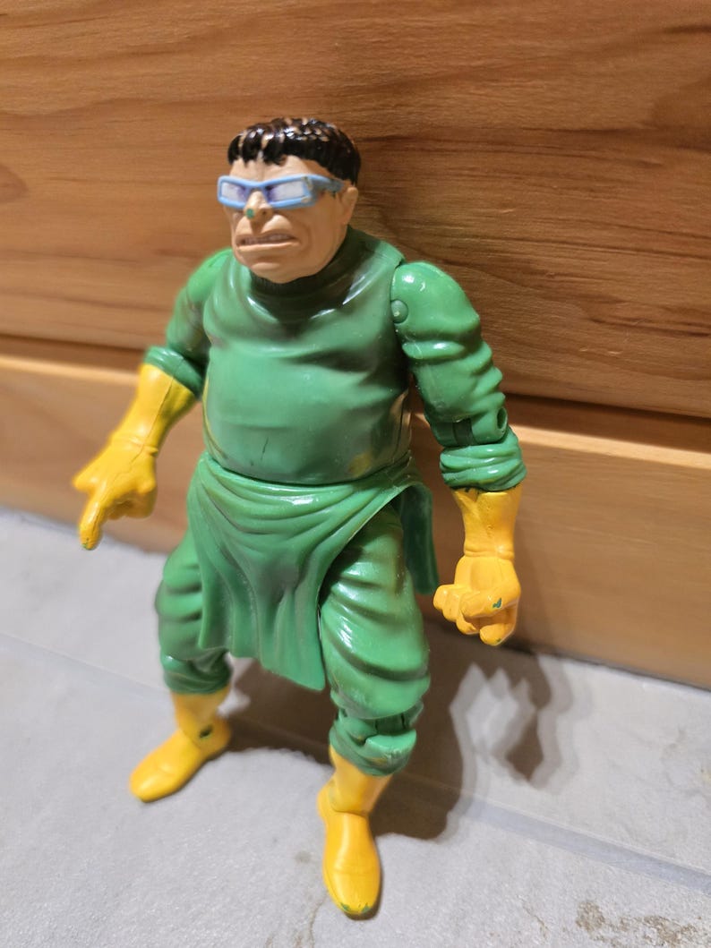 May include: A vintage action figure of a character in a green outfit with yellow gloves and boots. The figure has a stern expression, wearing blue-rimmed glasses. The figure is posed in a dynamic stance, suggesting action.