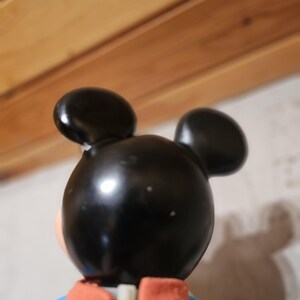 May include: A vintage Mickey Mouse figurine, viewed from the back. The figure has a black head with two round ears, a yellow jacket, a blue shirt, and red accents. The figurine is a collectible item.