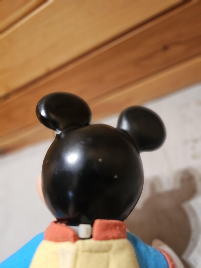 May include: A vintage Mickey Mouse figurine, viewed from the back. The figure has a black head with two round ears, a yellow jacket, a blue shirt, and red accents. The figurine is a collectible item.