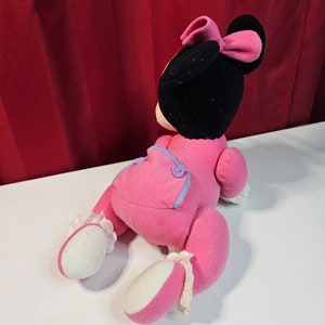 K&ouml;nnte beinhalten: Rosa Pl&uuml;schtier einer Comicmaus mit schwarzem Kopf und einer rosa Schleife. Das Spielzeug tr&auml;gt ein rosa-violettes Outfit mit wei&szlig;en Akzenten.